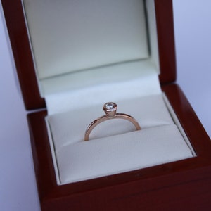 May include: A simple solitaire engagement ring with a single round diamond set in a rose gold band. The ring is displayed in a dark wood ring box with a white velvet interior.