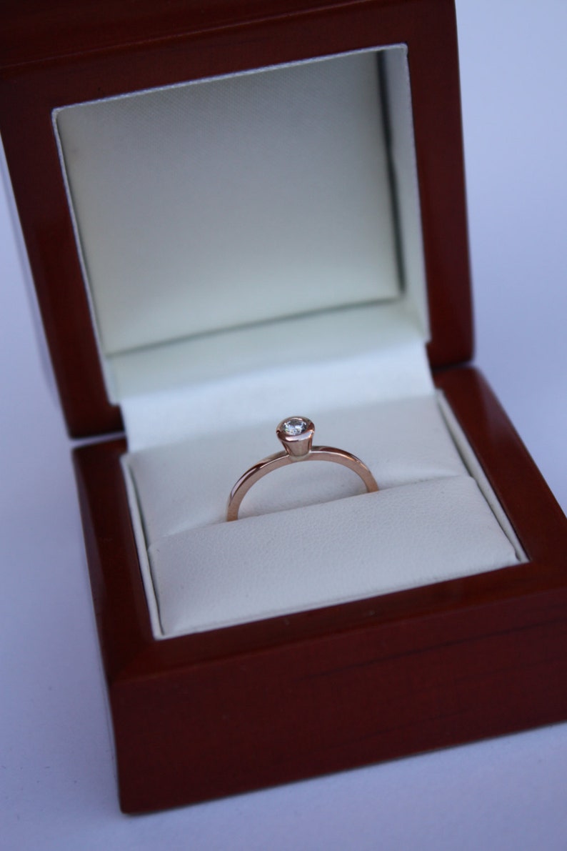 May include: A simple solitaire engagement ring with a single round diamond set in a rose gold band. The ring is displayed in a dark wood ring box with a white velvet interior.