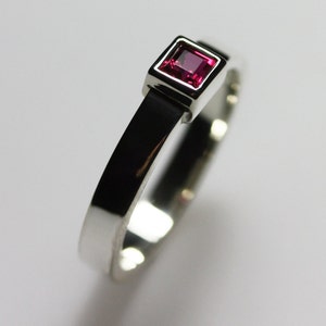 May include: A silver ring with a square, magenta-colored gemstone set in a bezel setting. The band is a simple, polished silver. The ring is shown on a white background.