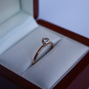 May include: A simple solitaire engagement ring with a single round diamond set in a rose gold band. The ring is displayed in a red velvet ring box.