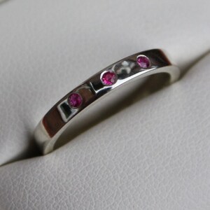 May include: A silver band ring with three small pink gemstones set in the band.