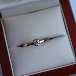 May include: A simple solitaire ring with a single round diamond set in a bezel setting. The ring is made of rose gold and is displayed in a wooden ring box.