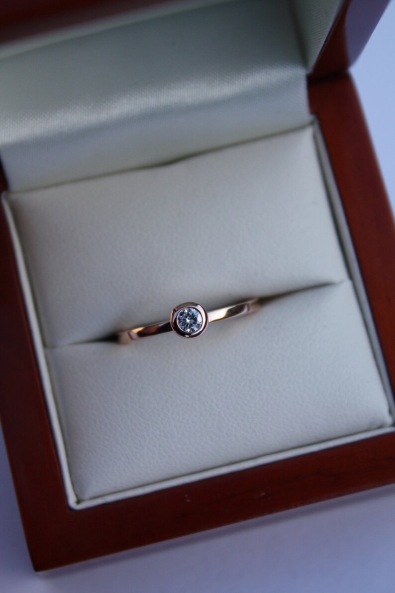 May include: A simple solitaire ring with a single round diamond set in a bezel setting. The ring is made of rose gold and is displayed in a wooden ring box.