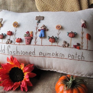 May include: A white pillow with embroidered details of a pumpkin patch, including pumpkins, sunflowers, and a scarecrow. The pillow has the text "old fashioned pumpkin patch" embroidered on it.