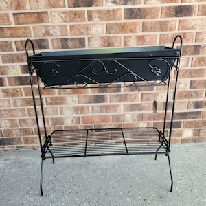 May include: Black metal plant stand with a rectangular planter box and a lower wire shelf. The planter box has decorative scrollwork and leaf accents. The stand is supported by slender legs and is suitable for indoor or outdoor use.
