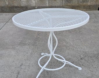 Op de afbeelding: Een witte ronde tafel met een cirkelvormig tafelblad met een metalen gaasoppervlak in een ruitpatroon. De tafel wordt ondersteund door een centrale voet die zich splitst in drie gebogen poten verbonden door een metalen ring.