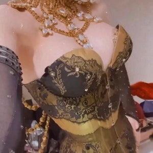 May include: A black and gold corset with intricate lace detailing and a sweetheart neckline. The corset is adorned with rhinestones and pearls.