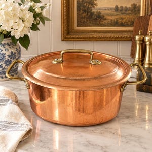May include: A round copper pot with a matching lid, featuring two brass side handles and a brass lid handle. The pot has a polished metallic finish and a smooth, rounded body. The design includes visible rivets securing the handles to the sides.