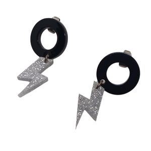 May include: A pair of dangle earrings featuring a black circular stud component attached to a silver-toned lightning bolt charm. The lightning bolt charms are covered in a fine silver glitter finish, and the earrings include standard metal post backs.
