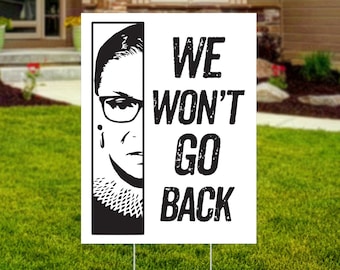 May include: Black and white yard sign with a portrait of Ruth Bader Ginsburg and the text "WE WON'T GO BACK".