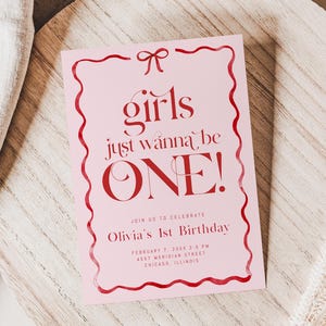 May include: A pink invitation card with red text and a decorative border. The text reads "girls just wanna be ONE!" and includes details for Olivia's 1st birthday. The card is on a wooden surface with a linen cloth and dried plant accents.