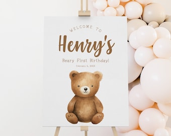 May include: Welcome to Henry's Beary First Birthday! A brown teddy bear sits on a white sign with brown text. The sign is on a wooden easel in front of a wall of beige balloons.