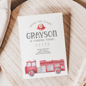 A birthday invitation with a red fire truck and a firefighter helmet in minimalist design. The text reads 'Sound the alarm! Grayson is turning three!