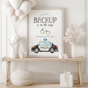 Puede incluir: Una impresión enmarcada con el texto "DADDY'S BACKUP is on the way!" con esposas y una ilustración de un coche de policía. La fecha de lanzamiento es julio de 2024. La obra de arte se exhibe sobre una mesa de madera con elementos decorativos.