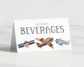 Puede incluir: Una tarjeta blanca plegada con el texto IN-FLIGHT BEVERAGES impreso en azul oscuro. Debajo del texto hay tres ilustraciones en acuarela de aviones: un biplano azul oscuro, un monoplano naranja y un avión de hélice azul claro.