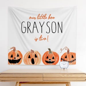 Op de afbeelding: Witte Halloween-banner met de tekst "our little boo GRAYSON is two!" in zwart en oranje. Vier waterverf pompoenen staan onderaan. Een kleine zilveren klok staat op een houten tafel.