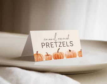 May include: A folded card on a white plate with the words "caramel covered PRETZELS" in black script and block letters. The card is decorated with watercolor pumpkins in shades of orange. The plate sits on a white cloth.