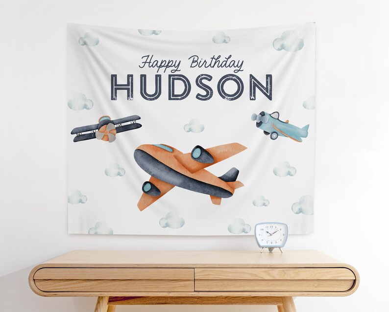 以下が含まれることがあります： 青い雲と「Happy Birthday Hudson」という文字が書かれた白い背景。オレンジとブルーの飛行機が2機あり、背景には白い雲がある。