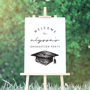 Pu&ograve; includere: Un cartello bianco su un cavalletto di legno con la scritta "WELCOME to alyssa's GRADUATION PARTY" in nero. Un disegno di un cappello di laurea &egrave; sotto il testo. Lo sfondo &egrave; un fogliame verde sfocato.