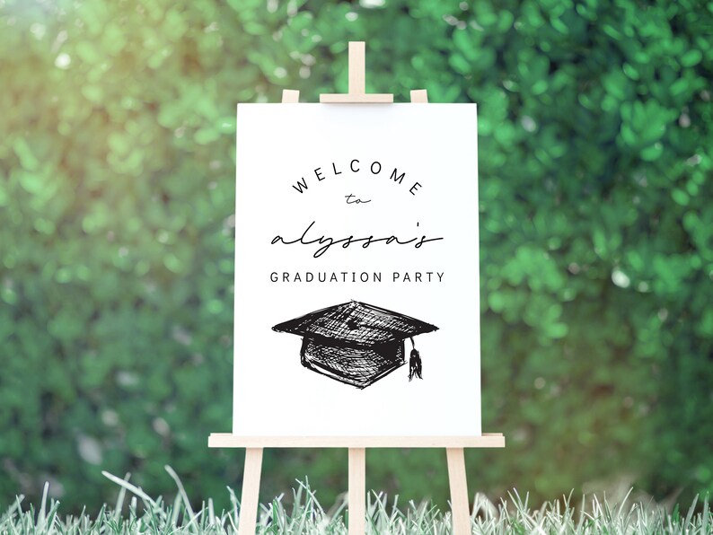 Pu&ograve; includere: Un cartello bianco su un cavalletto di legno con la scritta "WELCOME to alyssa's GRADUATION PARTY" in nero. Un disegno di un cappello di laurea &egrave; sotto il testo. Lo sfondo &egrave; un fogliame verde sfocato.