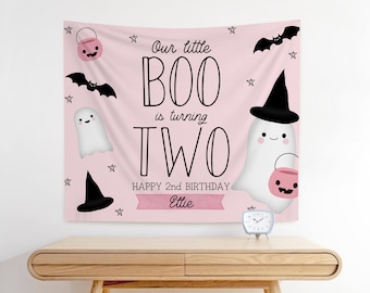 May include: A pink Halloween-themed banner with the text "Our little BOO is turning TWO" and "HAPPY 2nd BIRTHDAY Ettie". The banner features illustrations of ghosts, bats, stars, and jack-o'-lanterns. A small clock sits on a wooden table.