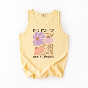 May include: A pale yellow sleeveless tank top featuring a graphic print of three stylized flowers in shades of purple and peach with brown stems. The text "BIG FAN OF HUMAN RIGHTS" is printed in a dark brown serif font above and below the flowers.