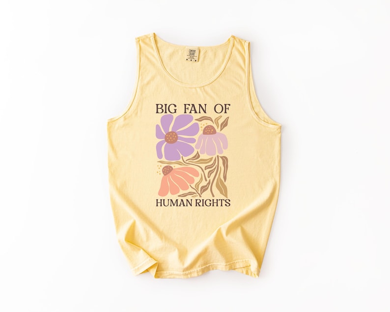 May include: A pale yellow sleeveless tank top featuring a graphic print of three stylized flowers in shades of purple and peach with brown stems. The text "BIG FAN OF HUMAN RIGHTS" is printed in a dark brown serif font above and below the flowers.
