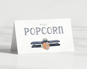 Puede incluir: Una tarjeta blanca con las palabras "PILOT POPCORN" en letras azul marino. Debajo, una ilustración en acuarela de un biplano con cuerpo azul y hélice naranja. La tarjeta está en ángulo.
