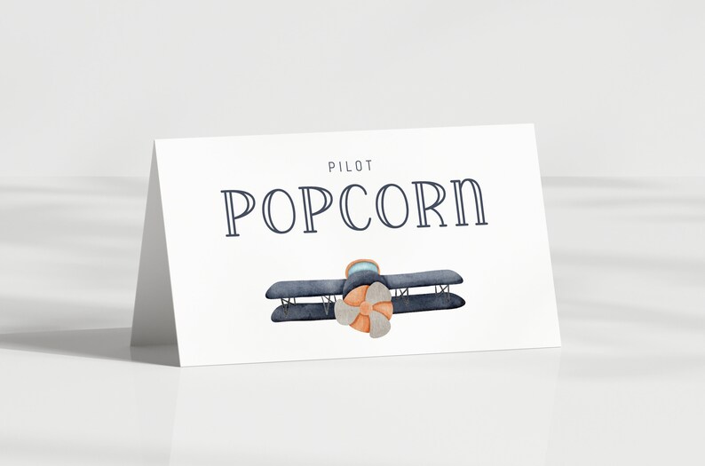 Może przedstawiać: Biała karta z napisem "PILOT POPCORN" w kolorze granatowym. Poniżej akwarelowa ilustracja dwupłatowca z niebieskim kadłubem i pomarańczowym śmigłem. Karta jest pochylona.