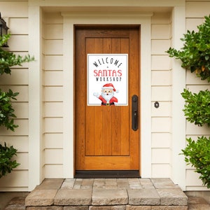 May include: A sign on a wooden door reads "WELCOME to SANTA'S WORKSHOP" in red and black text. A cartoon Santa Claus is featured below the text. The door is framed by white siding and green bushes.