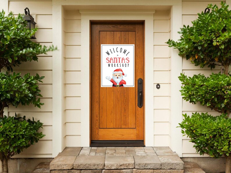 May include: A sign on a wooden door reads "WELCOME to SANTA'S WORKSHOP" in red and black text. A cartoon Santa Claus is featured below the text. The door is framed by white siding and green bushes.