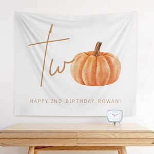 Puede incluir: Colgante de pared de tela blanca con una calabaza naranja y la palabra "two" en escritura marrón. El texto "HAPPY 2ND BIRTHDAY ROWAN!" está impreso debajo. Un pequeño reloj plateado está sobre una mesa de madera.
