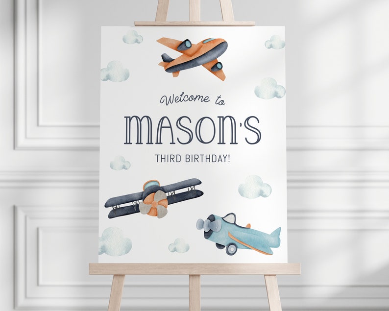 Może przedstawiać: Biały znak na sztaludze z akwarelowymi ilustracjami samolot&oacute;w i chmur. Znak głosi "Welcome to MASON'S THIRD BIRTHAY!" w granatowych i jasnoniebieskich literach. Samoloty są pomarańczowe, niebieskie i szare.