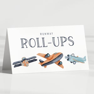 May include: A white folded card featuring the text RUNWAY ROLL-UPS in dark blue lettering. Below the text are three watercolor-style illustrations of airplanes: a dark blue biplane, an orange monoplane, and a light blue propeller airplane.
