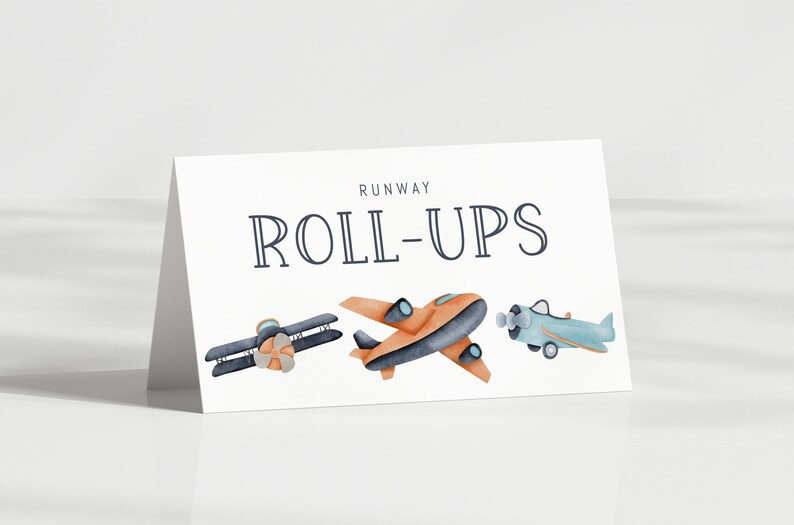 May include: A white folded card featuring the text RUNWAY ROLL-UPS in dark blue lettering. Below the text are three watercolor-style illustrations of airplanes: a dark blue biplane, an orange monoplane, and a light blue propeller airplane.