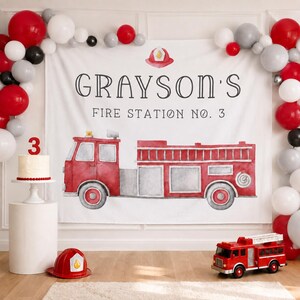 May include: A white fabric backdrop featuring a watercolor illustration of a red fire truck and the text "GRAYSON'S FIRE STATION NO. 3" with a small red fire helmet icon. The scene is decorated with a balloon arch in red, white, and gray.