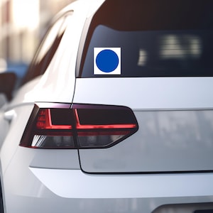 May include: A white car with a blue circle sticker on the rear window. The sticker is a solid blue circle inside a white square. The car's taillights are red, and the background is blurred.