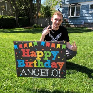 May include: A person is holding a black sign with colorful letters that say "Happy Birthday Angelo!" The sign has a colorful banner across the top and confetti on the background.