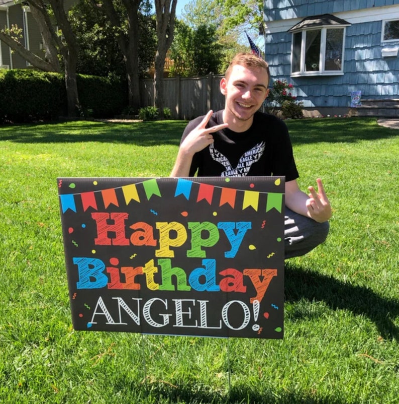 May include: A person is holding a black sign with colorful letters that say "Happy Birthday Angelo!" The sign has a colorful banner across the top and confetti on the background.