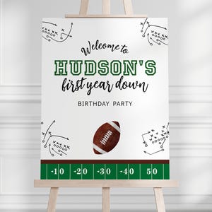 May include: White welcome sign with green and brown accents. The sign says "Welcome to HUDSON'S first year down BIRTHDAY PARTY". There is a football and football play diagrams on the sign.