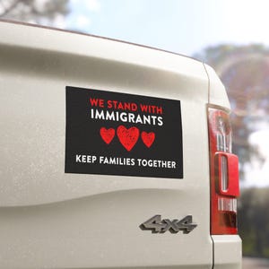 May include: A black rectangular bumper sticker with the text "WE STAND WITH IMMIGRANTS" in white, above three red hearts, and "KEEP FAMILIES TOGETHER" in white. The sticker is affixed to the back of a white vehicle.