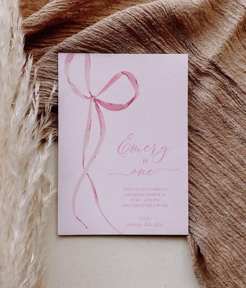 May include: Pink invitation with a watercolor illustration of a pink ribbon bow. The text reads "Emery is one" and includes details for a birthday party on Saturday, March 15th from 2:00 to 4:00 PM at 648 Cedar Hollow Rd. The invitation also includes the name Olivia and a phone number.