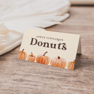 May include: A cream-colored card with the words "APPLE CINNAMON DONUTS" in dark brown lettering. The card features watercolor illustrations of orange pumpkins along the bottom. The card is on a wooden surface.