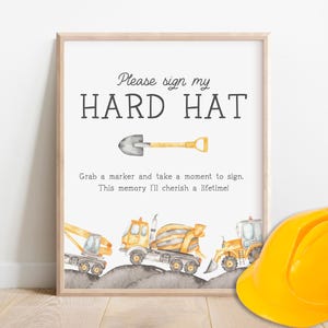May include: A printable sign with the text "Please sign my HARD HAT" and a shovel illustration. The sign is framed in wood and has a background of construction vehicles, including a cement mixer, a dump truck, and a backhoe.