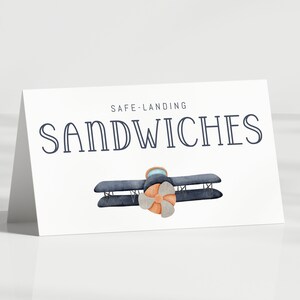 Może przedstawiać: Biała karta z napisem "SAFE LANDING SANDWICHES" w kolorze granatowym. Akwarelowa ilustracja dwupłatowca z brązowym śmigłem. Karta jest pochylona, rzucając cień.