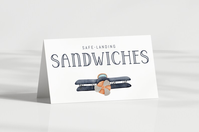 Może przedstawiać: Biała karta z napisem "SAFE LANDING SANDWICHES" w kolorze granatowym. Akwarelowa ilustracja dwupłatowca z brązowym śmigłem. Karta jest pochylona, rzucając cień.