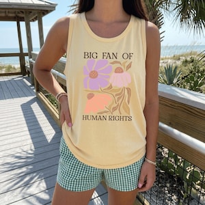 May include: A pale yellow sleeveless tank top featuring a graphic print of three stylized flowers in shades of purple, pink, and peach. The text "BIG FAN OF HUMAN RIGHTS" is printed in a dark brown serif font above and below the floral design.