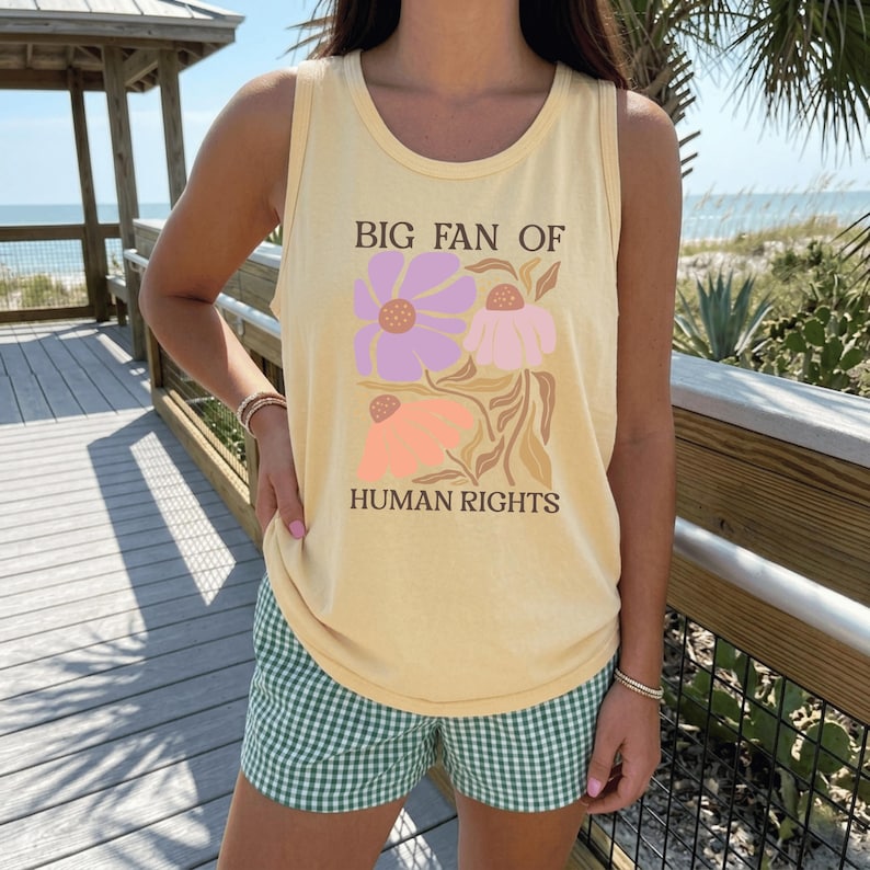 May include: A pale yellow sleeveless tank top featuring a graphic print of three stylized flowers in shades of purple, pink, and peach. The text "BIG FAN OF HUMAN RIGHTS" is printed in a dark brown serif font above and below the floral design.