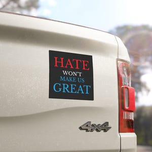 May include: A black rectangular bumper sticker with the words "HATE" in red, "WON'T MAKE US" in white, and "GREAT" in blue. The sticker is affixed to the back of a white vehicle.