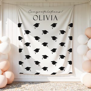 Op de afbeelding: Een witte afstudeerbanner met een patroon van zwarte afstudeerhoeden en de tekst "Congratulations! OLIVIA". De scène is versierd met witte en perzikkleurige ballonnen.